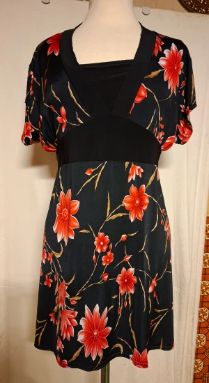 Svart blommig kortklänning från Tessa - Svart kortklänning från Tessa med stora röda och beige blommor, v-ringad hals och korta ärmar. Klänningen har ett svart midjeband med knyt baktill och ett mjukt, följsamt material som ger en skön passform. Perfekt för dig som gillar färgstarka prints och feminina detaljer.