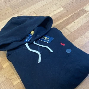 Svart hoodie från Polo Ralph Lauren - Svart hoodie från Polo Ralph Lauren med vit snörning och rött broderat logomärke på bröstet. Klassisk huva och långärmad modell i mjuk bomull. Storlek L