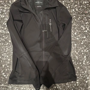 Svart vindjacka från Sail Racing - Svart vindjacka från Sail Racing med stilren design och diskret logga på bröstet. Jackan har dragkedja framtill, en bröstficka med dragkedja och två sidofickor. Materialet är syntet och jackan har lång ärm. Perfekt för dig som gillar sportig och clean look.