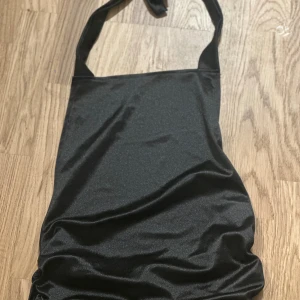 Svart halterneck topp  - Snygg svart topp med halterneck och slät, glansig yta. Toppen har en enkel och stilren design med knytband i nacken. Perfekt för dig som gillar minimalistisk stil och vill ha en topp som sticker ut med sin coola form.