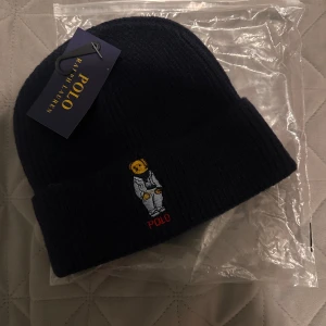 Mörkblå Polo Ralph Lauren mössa - Snygg mörkblå ribbstickad mössa från Polo Ralph Lauren med broderad astronaut och röd POLO-text framtill. Perfekt för dig som vill ha en cool accessoar med unik detalj. Materialet är mjukt och värmande.