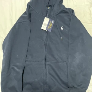Marinblå hoodie från Polo Ralph Lauren - Snygg marinblå hoodie från Polo Ralph Lauren med dragkedja och huva. Tröjan har långa ärmar, snörning vid huvan och klassisk logga broderad på bröstet. Perfekt för chill dagar och enkel att matcha med jeans eller joggers.