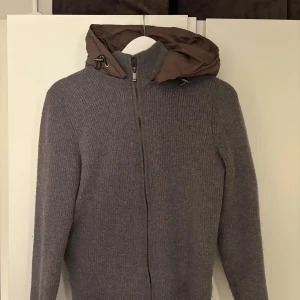 Grå stickad cardigan jacka med huva - Mysig grå stickad cardigan jacka från massimo dutti med dragkedja framtill och en brun quiltad huva. Jackan har långärmad design och ribbad struktur, perfekt för kyliga dagar. Enkel och stilren look som passar till det mesta.