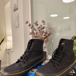 Svarta Dr. Martens Monet sneakers 36 - Svarta höga sneakers från Dr. Martens i textil med klassisk gul detaljsöm runt sulan. Skorna har snörning, rund tå och svart platt sula. Ikonisk Dr. Martens-flärp baktill och diskret logga på sulan. Perfekta för dig som gillar streetstyle och vill ha något bekvämt och snyggt.