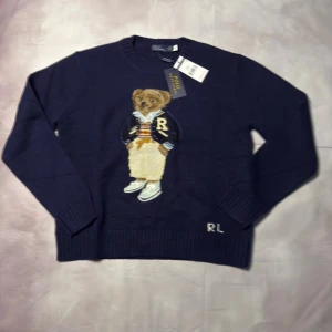 Mörkblå stickad tröja Polo Ralph Lauren - Mörkblå stickad tröja från Polo Ralph Lauren med ikonisk björn broderad på bröstet och RL-detalj vid nederkanten. Tröjan har rund hals, ribbade muddar och lång ärm. Materialet är 90% ull och 10% bomull, perfekt för att hålla dig varm och stilren.