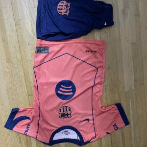 Barça fotbollströja Lamine Yamal #19 - Säljer en Barcelona fotbollströja med Lamine Yamal #19 på ryggen. Tröjan är korallrosa med mörkblå detaljer och tryck, inklusive klubbmärke och sponsorloggor. Kortärmad modell i lätt och ventilerande polyester, perfekt för match eller träning.