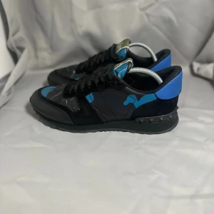 Valentino Rockrunner  - Snygga Valentino Rockrunner sneakers med svart och blått camouflagemönster. Skorna har svarta snören, platt sula med nitar och rund tå. Materialmix av skinn och mocka med blå detaljer på hälen och tungan. Perfekt för dig som gillar statement-sneakers.