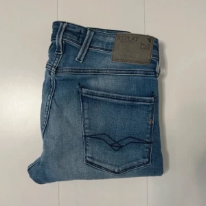 Blå Replay Anbass jeans herr - Snygga blå Replay Anbass jeans med klassisk femficksdesign och diskreta slitningar. Jeansen har normal passform, raka ben och tydliga sömmar på bakfickorna. Märkespatch i skinn baktill i midjan och detaljerad logga på myntfickan. Perfekta för en avslappnad stil.