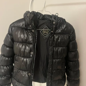 Svart superdry jacka  - Säljer en svart pufferjacka från XP-D i storlek 36. Jackan har en glansig finish, huva och dragkedja med tryckknappar. Den är vadderad, perfekt för kalla dagar. Inga skador