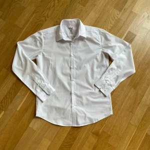 Vit skjorta från Smog till män, regular fit - Klassisk vit skjorta från Smog i storlek M med regular fit. Skjortan har lång ärm, knappar hela vägen framtill och klassisk krage. Tillverkad i bomull, enkel att matcha och har stilren look. Perfekt för dig som gillar clean och enkel stil.