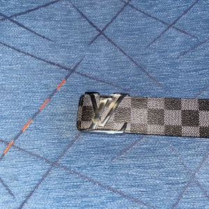 rutigt bälte i skinn - Säljer ett L bälte med ikonisk L-spänne i silver. Bältet har ett rutigt mönster i svart och grått på framsidan och är tillverkat i skinn. Baksidan är beige med synliga sömmar. Perfekt accessoar för dig som gillar designer och vill sticka ut.