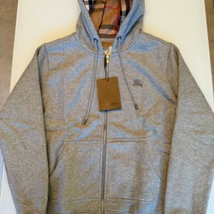 Grå hoodie från Burberry med dragkedja - Grå hoodie från Burberry med klassiskt rutigt foder i huva och insida. Tröjan har dragkedja framtill, snörning i huvan och två fickor. Tillverkad i mjuk bomullsmix och har diskret Burberry-logga på bröstet. Perfekt för en stilren och avslappnad look.