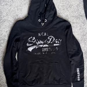 Svart Superdry hoodie med tryck - Svart hoodie från Superdry med stort logotryck och japanska tecken på bröstet och ärmen. Tröjan har huva med snörning och en klassisk magficka framtill. Perfekt för dig som gillar streetwear och vill ha en cool look.