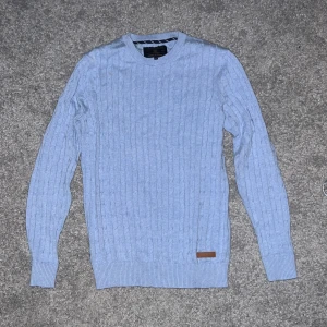 Ljusblå Bondelid stickad tröja S - Snygg ljusblå stickad tröja från Bondelid i storlek S. Tröjan har ett klassiskt kabelstickat mönster, rund hals och ribbade muddar vid ärmslut och nederkant. Liten Bondelid-logga i brunt vid nederkanten ger en schysst detalj. Perfekt för dig som gillar stilren och enkel design.