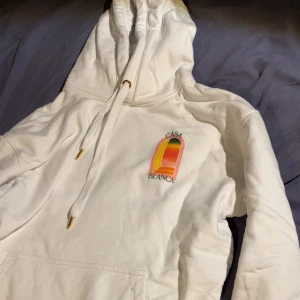 Vit hoodie från Casablanca med tryck - Säljer en vit hoodie från Casablanca med färgglatt grafiskt tryck fram och bak. Tröjan har huva med snörning och guldfärgade detaljer på snöret. Mjuk bomullskvalitet och klassisk känguruficka framtill. Perfekt för dig som gillar streetwear och snygga prints.