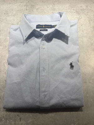 Ljusblå skjorta från Ralph Lauren - Snygg ljusblå skjorta från Ralph Lauren i klassisk passform. Skjortan har knappar framtill och den ikoniska broderade loggan på bröstet. Tillverkad i mjuk bomull, perfekt för en clean och stilren look. Krage med knappar och diskret design.