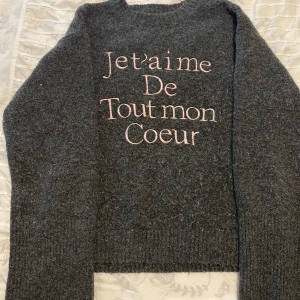 Mörkgrå stickad tröja med broderi - Mysig mörkgrå stickad tröja med rosa broderad text 'Je t'aime de tout mon coeur' på framsidan. Tröjan har långärmade ribbstickade muddar och en klassisk rund hals. Perfekt för dig som gillar franska vibes och vill ha något extra mjukt och varmt.
