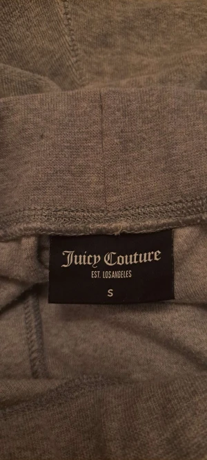 Grå mjukisbyxor Juicy Couture S - Grå mjukisbyxor från Juicy Couture i storlek S. Byxorna har två bakfickor med silverfärgade knappar och broderad JC-logga. Materialet är mjukt och skönt, perfekt för chill dagar. Klassisk rak passform med normal midja.