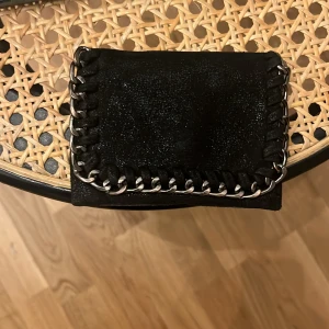 Svart plånbok med kedjedetaljer - Snygg svart plånbok i skinn med coola silverfärgade kedjedetaljer runt kanterna. Plånboken har flera fack för kort och stängs med tryckknapp. Perfekt accessoar för dig som gillar en edgy och stilren look.
