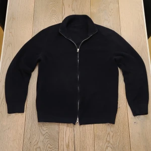 Mörkblå stickad kofta med dragkedja - Mörkblå stickad kofta med hel dragkedja och hög krage. Snygg och enkel design som funkar till jeans eller chinos. Långärmad och med ribbade muddar för extra komfort. Perfekt för dig som gillar stilrena plagg med lite edge. Vet tyvärr varken vilket märke den är eller vilken storlek den är men skulle tippa på att den är stl S då den passar mig som brukar ha det. Den är mörkblå, inte svart men ljuset får den att se mörkare ut