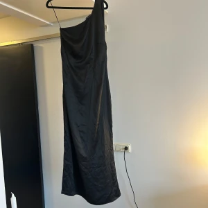 Svart långklänning från Adoore - Snygg svart långklänning från Adoore i slätt, glansigt material. Endast använd en gång. Klänningen har en oneshoulder-design med ett smalt axelband och en elegant drapering över axeln. Passformen är normal och klänningen har en stilren, minimalistisk look som känns modern och chic.