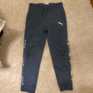 Mörkblå mjukisbyxor från Puma - Snygga mörkblå mjukisbyxor från Puma med elastisk midja och snörning. Byxorna har coola mönstrade detaljer längs benen och ett vitt Puma-logotryck på framsidan. Perfekta för chill eller träning, med ribbade muddar vid benslut.