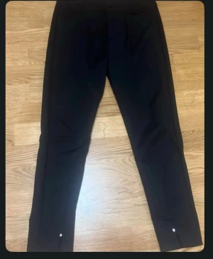 Svarta kostymbyxor med smal passform - Snygga svarta kostymbyxor med smal passform och raka ben. Byxorna har diskreta knappar vid bensluten och klassisk design som passar till skjorta eller t-shirt. Perfekta för dig som gillar stilrena och enkla plagg.