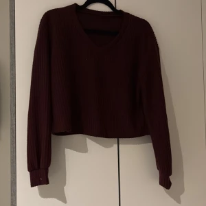 Vinröd ribbad croppad tröja XS - Snygg vinröd croppad tröja med ribbad struktur och v-ringad hals. Långärmad modell som passar perfekt till jeans eller kjol. Mjuk och skön, tillverkad i ett stickat material. Enkel att styla för en chill och trendig look.