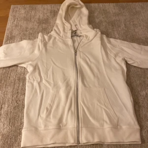 Vit hoodie med dragkedja och huva - Vit hoodie med hel dragkedja framtill och stor huva. Tröjan har två fickor på magen och ribbade muddar vid ärmslut och nederkant. Perfekt för chill dagar och enkel att matcha med jeans eller mjukisbyxor.