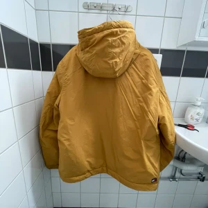 Beige vindjacka från Tommy Hilfiger - Säljer en beige vindjacka från Tommy Hilfiger med huva och klassisk logga på bröstet. Jackan har långa ärmar, tryckknappar framtill och två stora fickor. Perfekt för dig som gillar streetwear och vill ha en chill look.