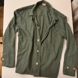 Grön overshirt från Armor Lux - Snygg grön overshirt från franska Armor Lux med klassisk krage och stora knappar framtill. Skjortan har två stora fickor framtill och är tillverkad i ett slitstarkt bomullstyg. Perfekt lager-på-lager-plagg med lång ärm och enkel, stilren design. Endast använd ett fåtal gånger.