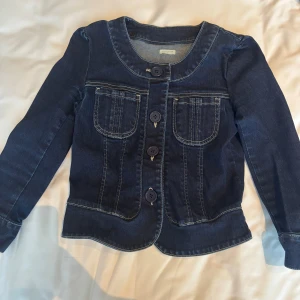 Mörkblå jeansjacka från PersoJea - Snygg mörkblå jeansjacka från PersoJea med rund halsringning, kontrastsömmar och stora knappar framtill. Jackan har två bröstfickor och är långärmad. Perfekt för dig som gillar klassisk denimstil med en modern twist.