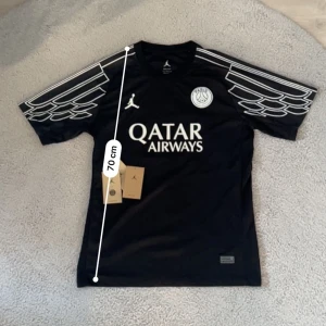 Svart PSG x Jordan fotbollströja - Snygg svart PSG fotbollströja i samarbete med Jordan, med vita detaljer och tryck. Tröjan har korta ärmar med unikt vitt mönster på axlarna, rund hals och tryckta loggor för Qatar Airways och PSG. Tillverkad i lätt och ventilerande polyester.Aktiv 24/7✅ Skickas inom 24h📦🚚 Kan gå ner i hugget 😃✅ Hör av er via frågor⁉️