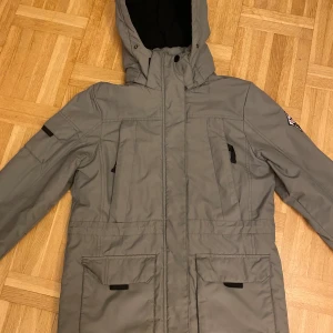 Vinterjacka - Grå parkajacka från Cubus med huva och cool North Pole Adventure-patch på ärmen. Jackan har flera fickor med lock, svarta detaljer och quiltad insida. Materialet är polyamid och polyester, perfekt för kalla dagar. Snygg och funktionell design med dragkedja framtill. PRISET KAN DISKUTERAS.