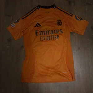 Real Madrid Mbappé fotbollströja - Helt ny // Snygg orange Real Madrid fotbollströja från Adidas med Mbappé och nummer 9 på ryggen. Tröjan har svarta detaljer, klubbmärke på bröstet och sponsortryck framtill. Korta ärmar och tillverkad i lätt, ventilerande material. Perfekt för dig som älskar fotboll och vill sticka ut på planen.