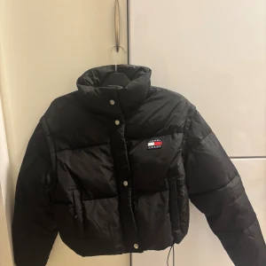Svart pufferjacka från Tommy Jeans - Svart Tommy Jeans pufferjacka med avtagbara ärmar som gör den omvandlingsbar till väst. Faux dun-fylld för värme, hög krage och dragkedja + tryckknappar framtill. Två hip-fickor, resår vid ärmsluten och logga på bröstet. Tillverkad av återvunnet material – perfekt för kalla dagar och streetwear!