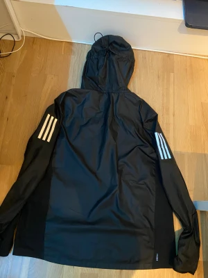 Svart vindjacka från Adidas med huva - Svart vindjacka från Adidas med huva och vita ränder på båda ärmarna. Perfekt för löpning med sportig look och RUNNING-detalj på insidan. Vill bli av med jackan, priset går såklart att diskutera. :) 