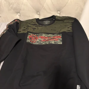 Philipp Plein svart tröja med camo-detalj - Svart långärmad tröja från Philipp Plein med coola militärgröna och camouflage-detaljer över bröstet och axlarna. Röd broderad logga framtill. Unik design med slitna partier upptill och camo-tryck. Perfekt för dig som gillar streetwear med edge.