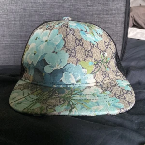 Gucci blommig keps med mesh - Snygg keps från Gucci med beige och svart GG-mönster, täckt av blå och turkosa blommor. Framsidan är i canvas med blommigt tryck, medan baksidan är i svart mesh för extra ventilation. Justerbar snapback och böjd skärm. Perfekt för dig som vill sticka ut. Säljer denna keps för den har aldrig kommit till användning 