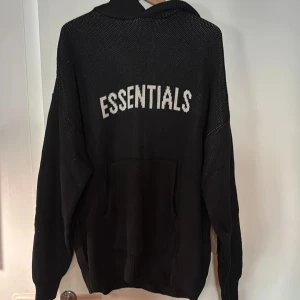Svart Essentials hoodie stickad XL - Stickad hoodie från Fear of God Essentials i beige med svart logga på ryggen. Oversized fit, huva och känguruficka framtill. Perfekt för dig som gillar streetwear och vill ha en chill vibe. XL men väldigt stor i storleken 