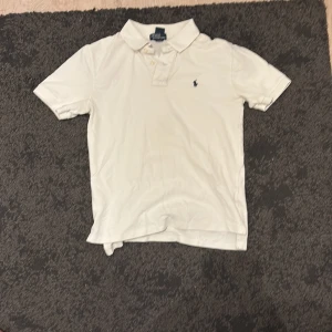 Vit pikétröja från Polo Ralph Lauren - Klassisk vit pikétröja från Polo Ralph Lauren med korta ärmar, krage och knappar framtill. Tröjan har den ikoniska broderade loggan på bröstet och är tillverkad i bomull för en skön och luftig känsla.