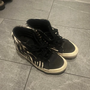 Vans zebra-mönstrade high top sneakers - Säljer ett par Vans high top sneakers med svart och vitt zebramönster i pälsimitation på sidorna. Skorna har svarta detaljer, snörning och vit sula. Klassisk Vans-logga baktill och ikonisk vågig sidostrip. Perfekt för dig som vill sticka ut med coola detaljer.