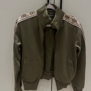 Olivgrön bomberjacka från Guess - Säljer en olivgrön bomberjacka från Guess med beige axelband och svarta loggor längs axlarna. Jackan har ribbade muddar, dragkedja och två fickor framtill. Perfekt för dig som gillar streetstyle och vill ha en snygg jacka med coola detaljer.
