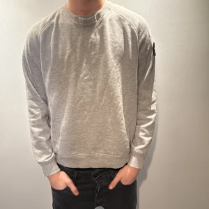 Grå sweatshirt från Stone Island - Säljer en grå sweatshirt från Stone Island med klassisk rund hals och ribbade muddar. Tröjan har den ikoniska Stone Island-loggan på vänster ärm och är tillverkad i mjuk bomull. Perfekt för dig som gillar stilren streetwear. Modellen är 186cm och väger 70kg 