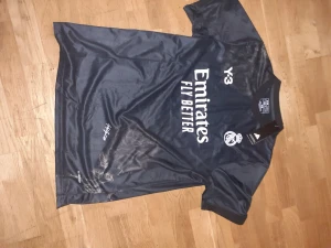 Real Madrid Y-3 x Adidas fotbollströja - Snygg svart Real Madrid fotbollströja från Adidas och Y-3, med vit logga och tryck på bröstet. Tröjan har korta ärmar, rund hals och är gjord i lätt, ventilerande polyester. Perfekt för dig som vill sticka ut på planen eller på läktaren.