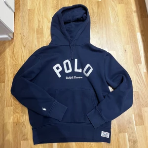Marinblå hoodie från Polo Ralph Lauren - Säljer en marinblå hoodie från Polo Ralph Lauren med stor vit POLO-text och broderad logga på ärmen. Klassisk känguruficka, huva med snörning och mjukt material. Perfekt för dig som gillar stilren streetwear.                                                               Nypris: 2500kr