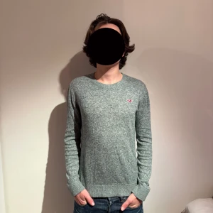 Grå stickad tröja från Hollister  - Snygg gråmelerad stickad tröja från Hollister i storlek XS. Tröjan har rund halsringning, långa ärmar och en liten röd Hollister-logga broderad på bröstet. Perfekt för lager-på-lager och chill vardagsstil. Modell 175cm 60kg