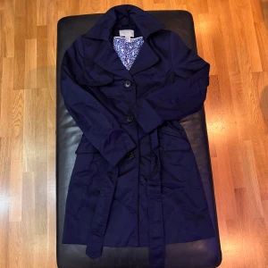Mörkblå trenchcoat från H&M, strl 34 - Snygg mörkblå trenchcoat från H&M i storlek 34. Jackan har klassisk krage, knappar framtill och bälte i midjan. Fodret har ett blåvitt grafiskt mönster. Perfekt för dig som vill ha en stilren och tidlös look.
