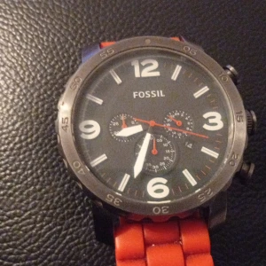 Fossil klocka med orange silikonband - Snygg och sportig Fossil klocka med stor rund svart urtavla och tydliga vita siffror. Klockan har tre små kronografer och ett coolt orange silikonarmband som ger en modern vibe. Metallboett i mörkgrått och flera knappar på sidan för extra funktioner.