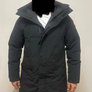 Svart dunjacka från Canada Goose - Svart dunjacka från Canada Goose med stor huva och klassisk logga på ärmen. Jackan har två stora fickor framtill och dragkedja med vindslå. Perfekt för kalla dagar och har en clean, stilren look som funkar till allt. Såklart äkta.